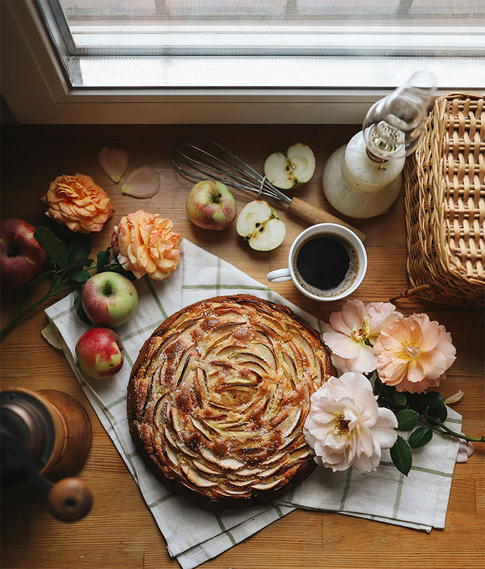 Beautiful photo of an apple pie cooling under a rustic window, surrounded by a hurricane oil lamp, an old-fashioned whisk, apple slices, camellia flowers, and a fresh cup of coffee